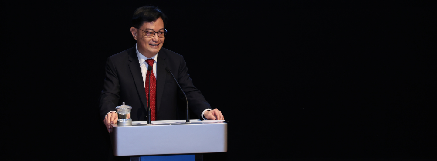Man with glasses in suit & red tie stands at lectern. Water pitcher visible, black background.